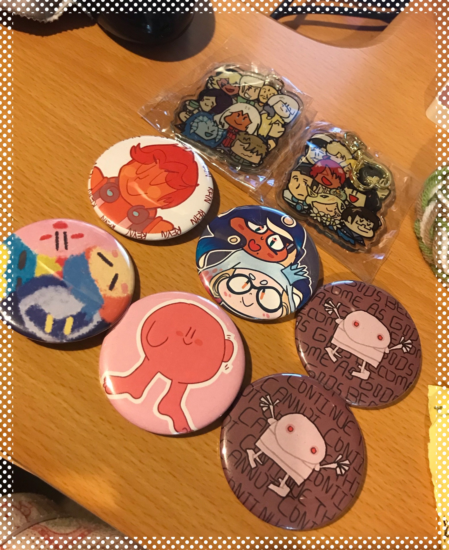 photo of assorted buttons and acrylic charms. from top to bottom left to right, is a xenoblade x group charm front and back, then a reyn xenoblade pin, off the hook splatoon pin, nier automata robot become as gods pin, kirby's return to dreamland pin, kirby with feet pin, and nier automata robot this cannot continue pin. they are displayed on a wooden desk with a polka dot frame around the pic.